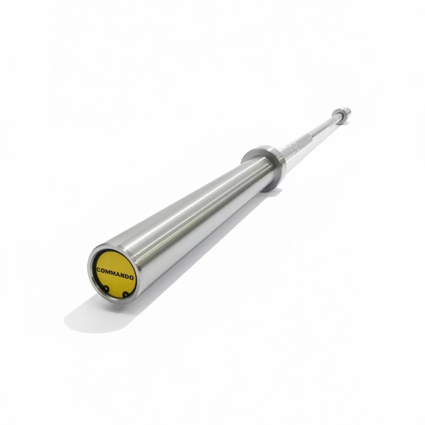 A silver metal barbell with a yellow Commando logo on the end cap, resting on a black gym floor with exercise equipment in the background.