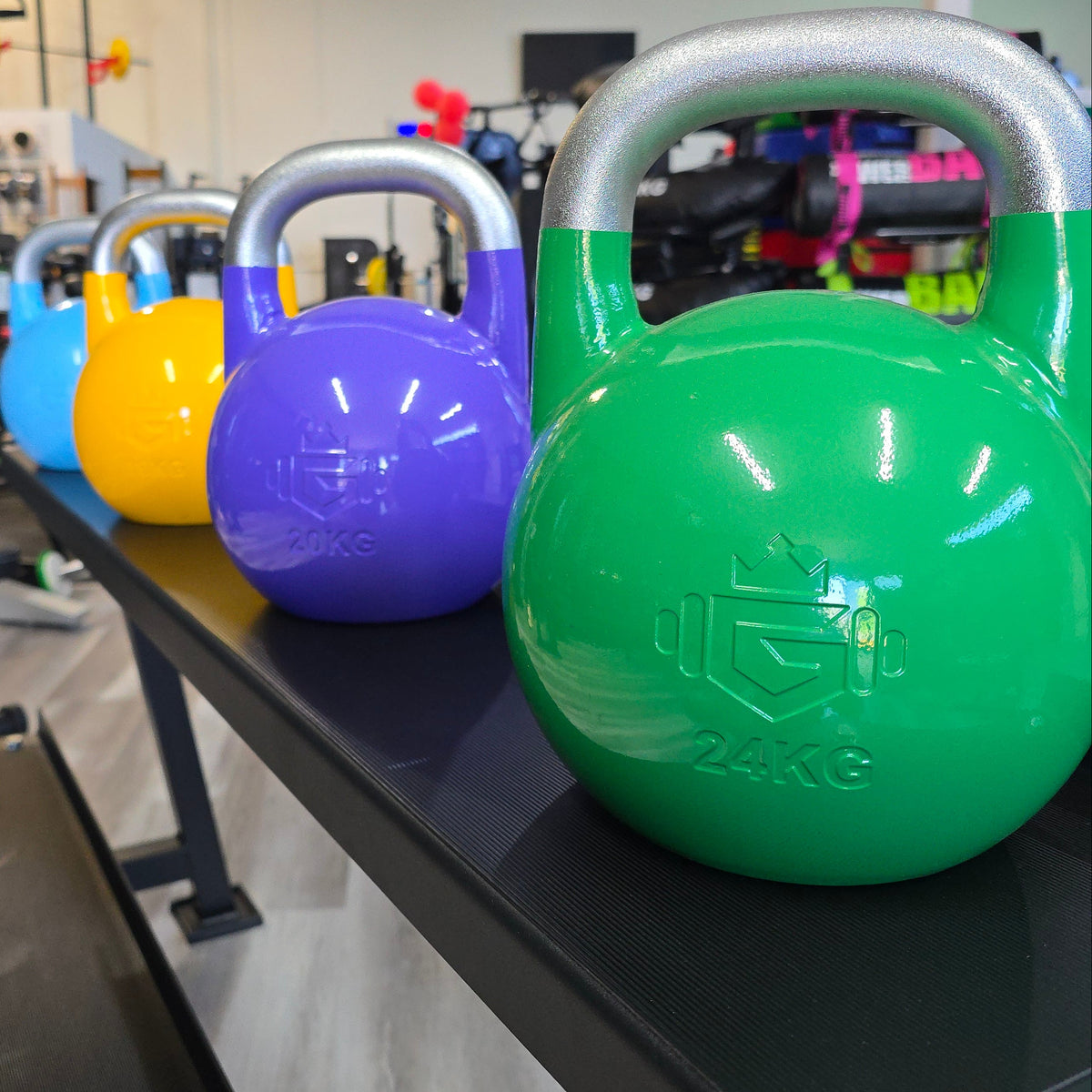 A row of colorful 24kg kettlebells in green, purple, yellow, and blue, displayed on a black gym bench inside a fitness center.