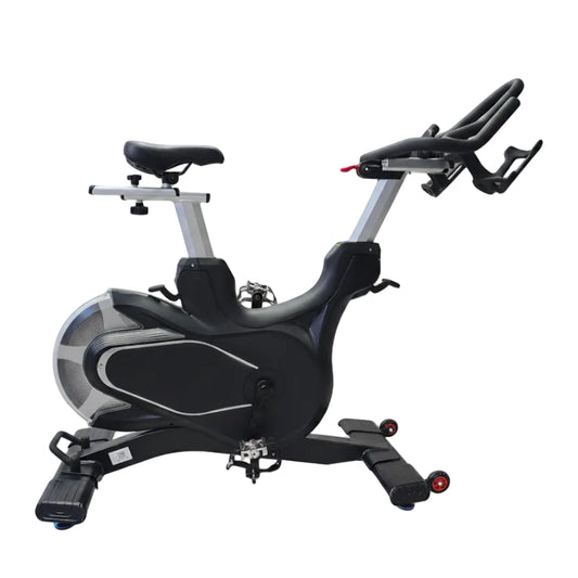 A black and silver indoor stationary exercise bike with a black seat, padded handlebars, and a flywheel, photographed indoors against a white draped backdrop on a light wood floor.