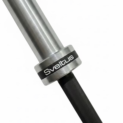 Close-up of a metal barbell with a black grip and a black collar ring featuring the white Sveltus brand name, resting on a dark textured gym floor.