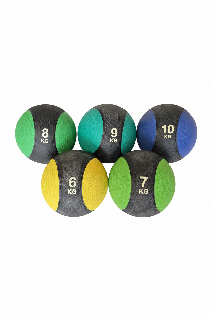 Five medicine balls in different weights (6 kg to 10 kg) with black, green, blue, yellow, and teal panels, displayed on a black mat with stacked cardboard boxes in the background.