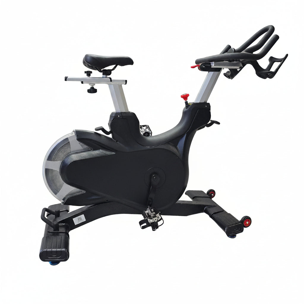 A black and silver indoor stationary exercise bike with the word 'FITNESS' printed on the side, photographed indoors against a large white fabric backdrop on a light wood floor.