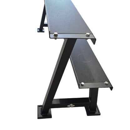 A black metal workout table with two integrated benches and square base plates, photographed in a gym with wood-look flooring and exercise equipment in the background.