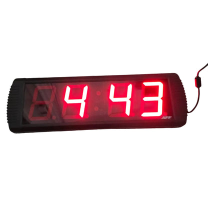 A rectangular digital LED countdown timer showing 4:43 in bright red numbers, placed on a gray wood-look floor against a white wall with visible power cords and outlets.