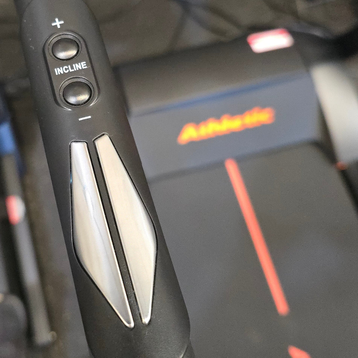 Close-up of a treadmill console handlebar with speed and incline buttons, showing a black textured grip with silver accents, with another treadmill in the background.